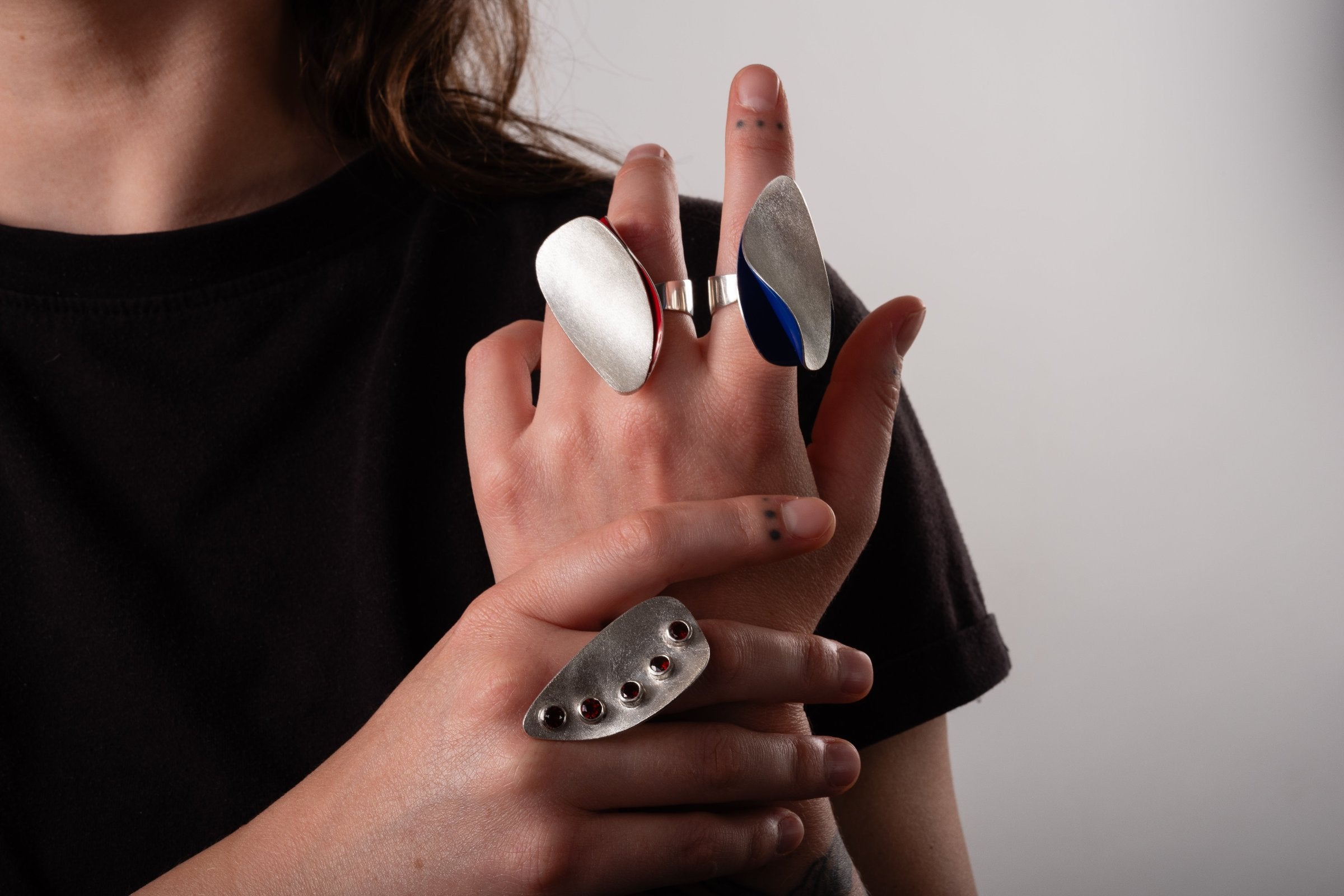 Three big statement silver rings worn on the hands of a woman.  Two rings have coloured enamel inside in red and in blue.  The third ring has five large red garnets.