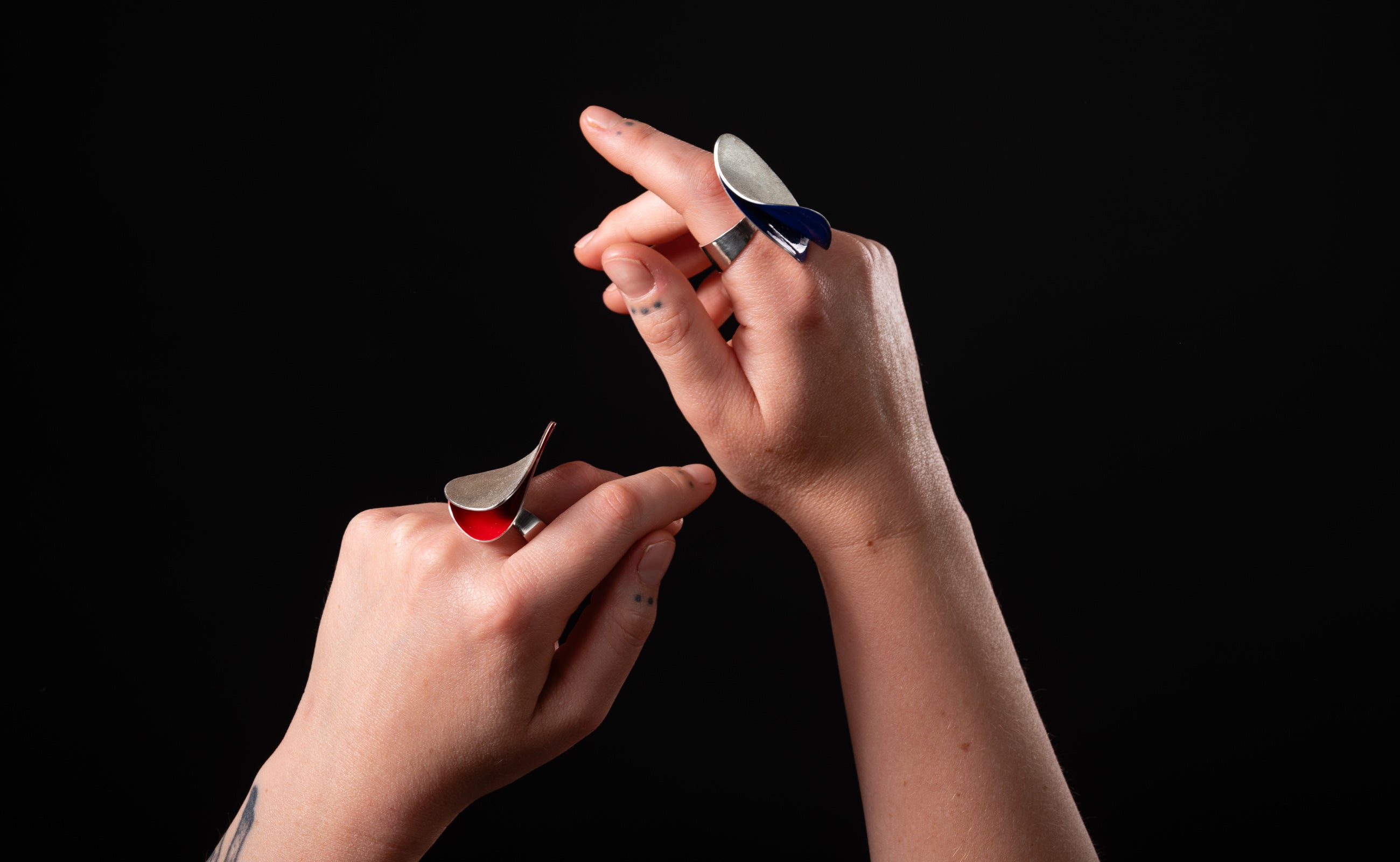 Two hands each wearing a large sculptural silver ring.  One ring has red enamel peaking out and the other ring has blue enamel.