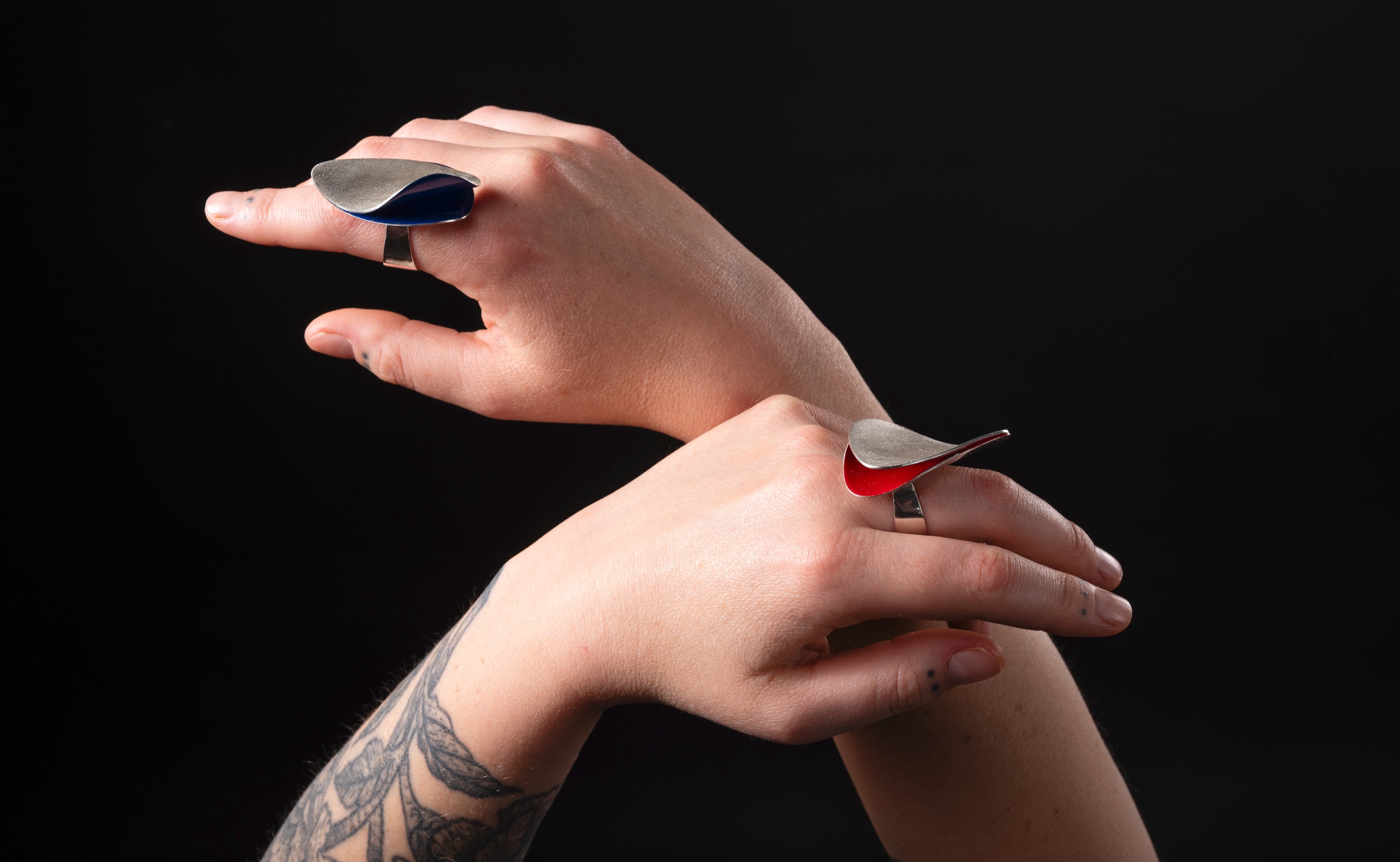 A woman's hands held up and each wearing a bold silver ring.  Each ring has a curved shape and frosted texture with bright colourful enamel revealed in blue on one ring and red on the other.