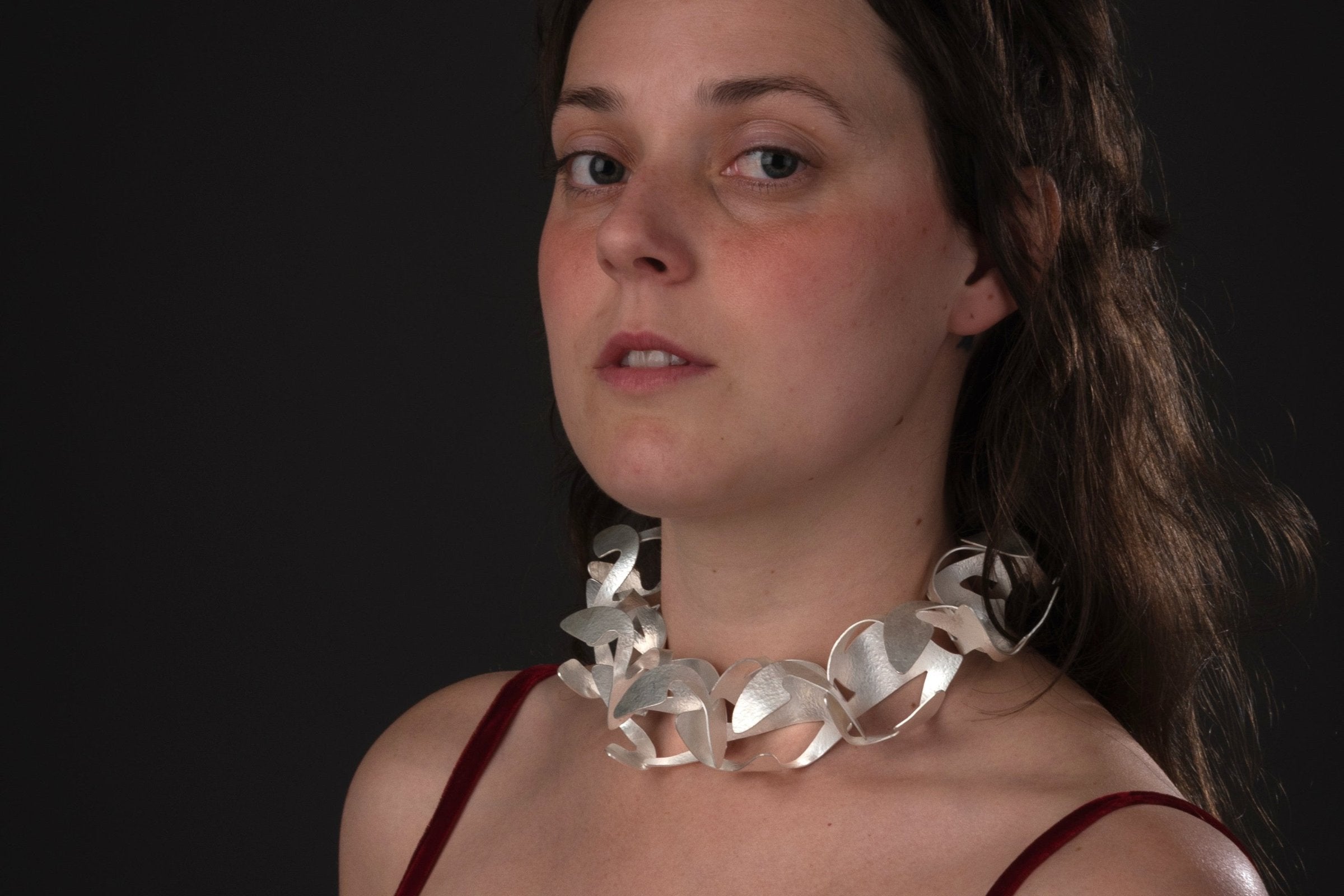 A woman in a red velvet evening dress wearing a large statement silver collar necklace made up of repeating organic shaped forms.